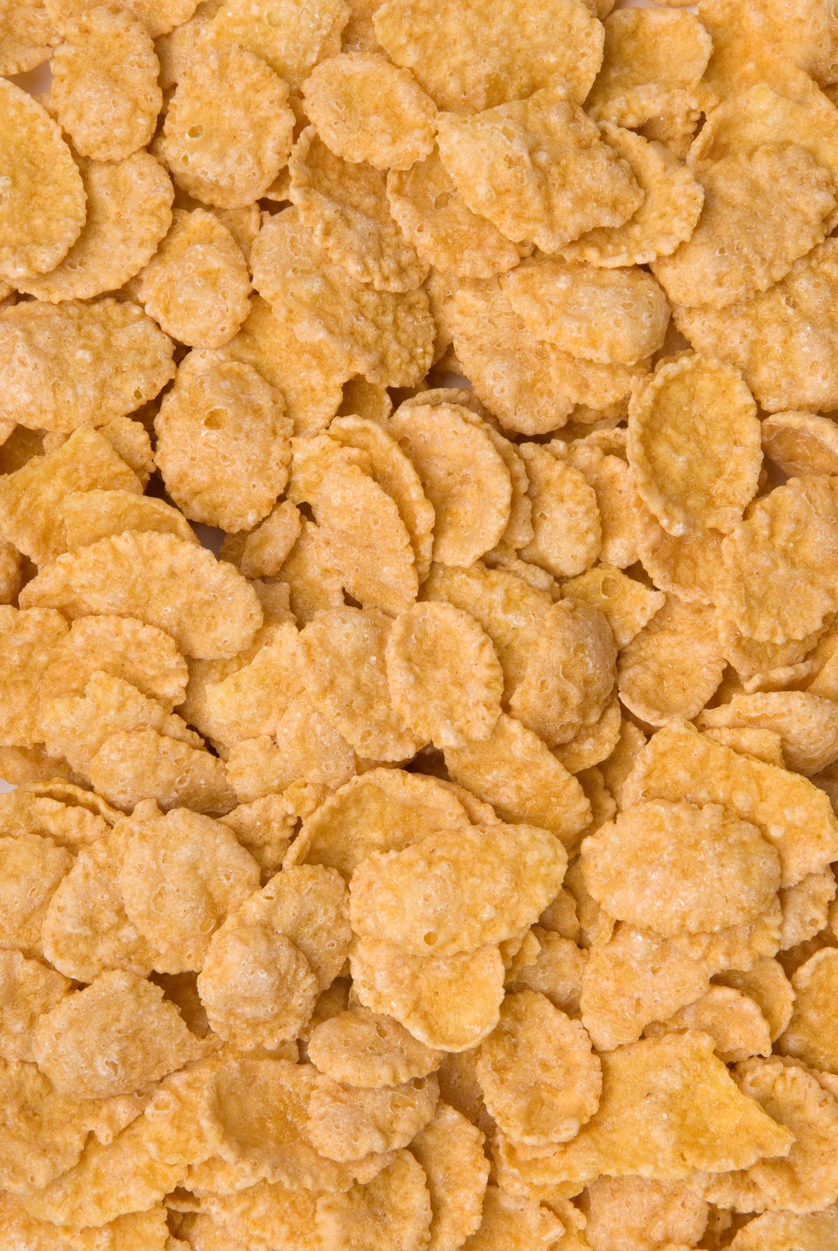 A close-up view of plain golden cornflakes, showing their rough texture and irregular shapes, piled together to fill the entire image.