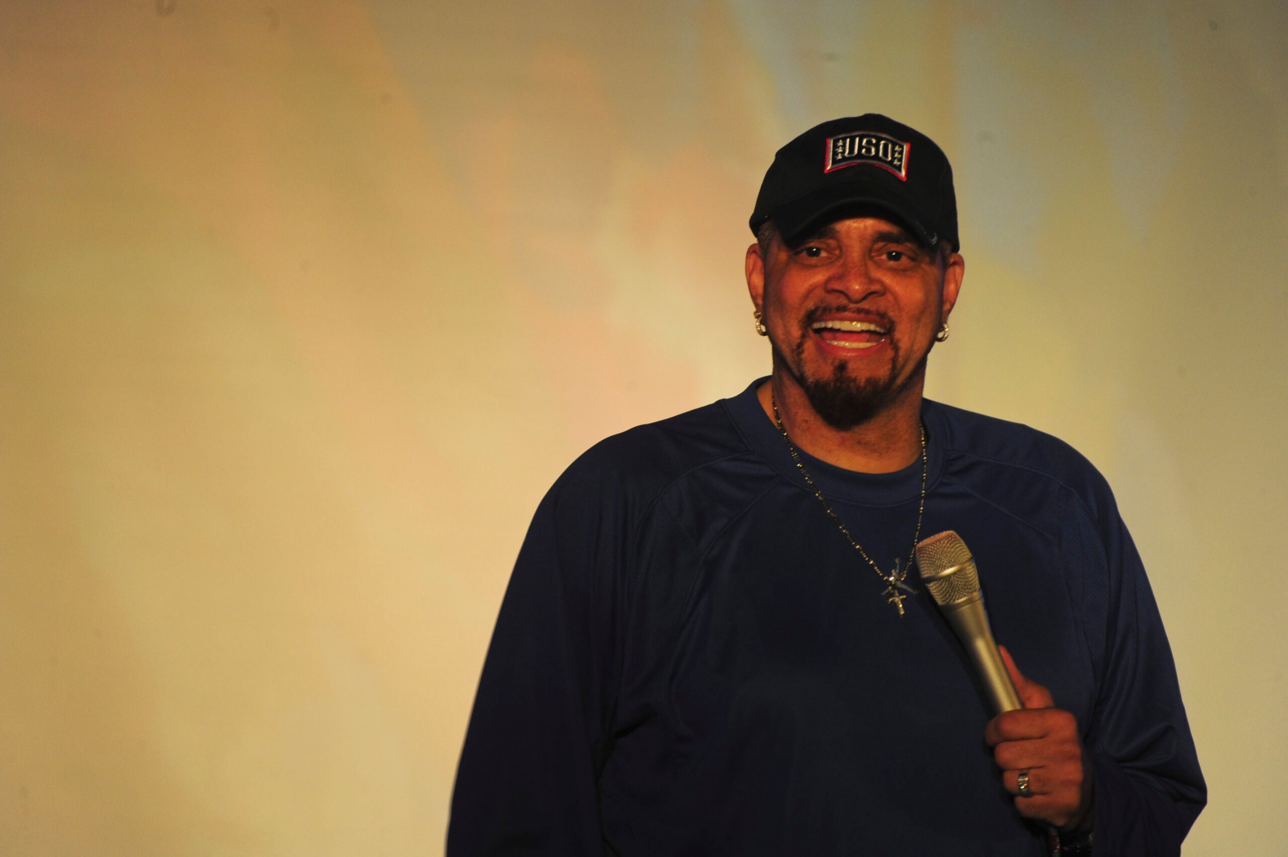 A man wearing a black cap, dark blue shirt, and a chain necklace is smiling while holding a microphone, standing against a blurred, light-colored background.