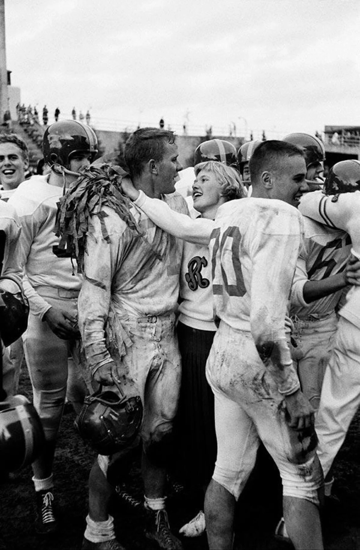 A smiling cheerleader hugs a muddy football player while surrounded by teammates celebrating on the field after a game.