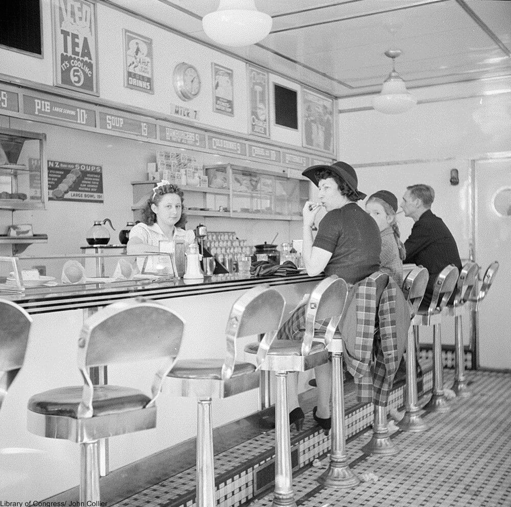 Black and white photo of people sitting at a diner counter, with a waitress behind the counter. Vintage posters and a menu hang on the wall above. The floor has a checkered pattern, and shiny stools line the counter.
