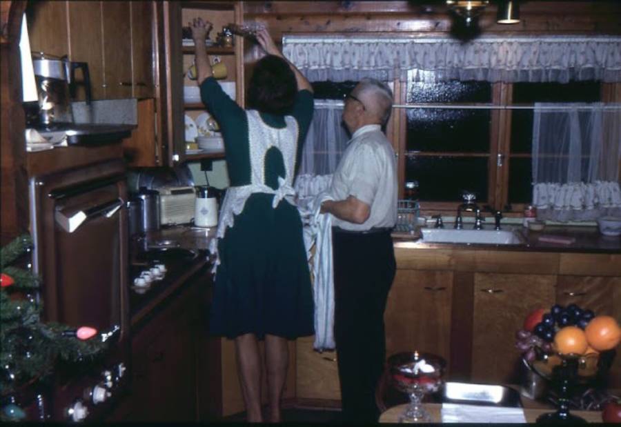 A woman in a white apron reaches for a shelf in a cozy, wood-paneled kitchen while an older man stands nearby holding a dish towel. Fruit and dishes are on the counter.