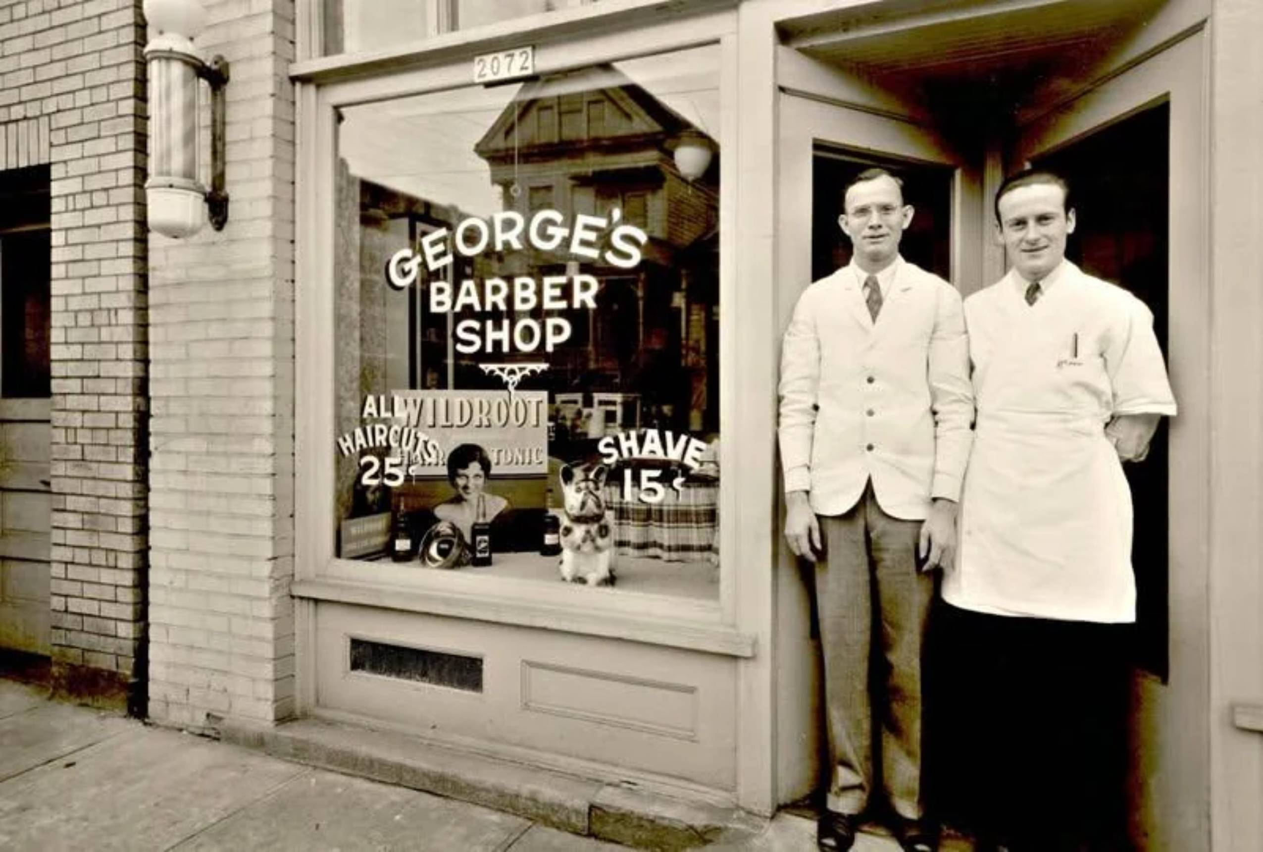 Two barbers stand outside George’s Barber Shop. The window advertises haircuts for 25 cents and shaves for 15 cents. A dog and a child are visible inside by the window. The setting appears vintage, circa early 20th century.