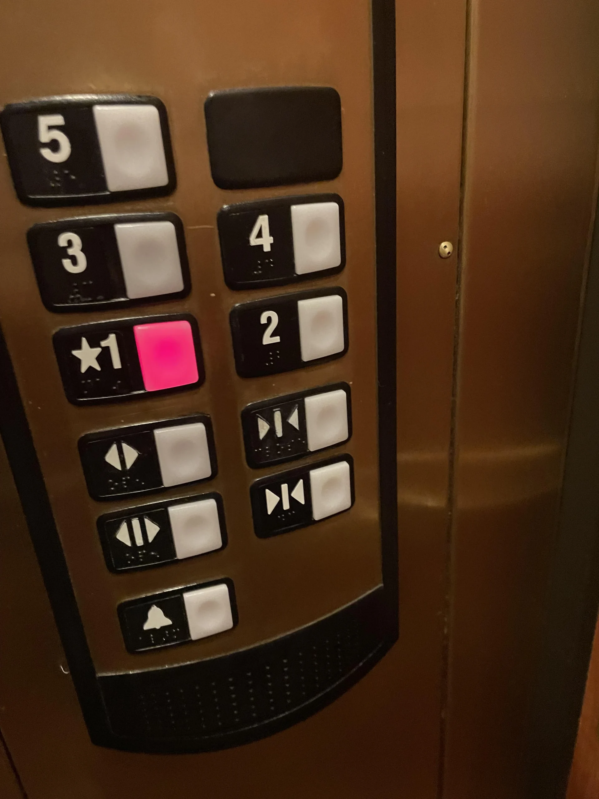 Close-up of an elevator control panel with buttons labeled 1 to 5 and door controls. The button for floor 1 is lit red, indicating it has been selected. The panel is metallic with black, white, and red elements.