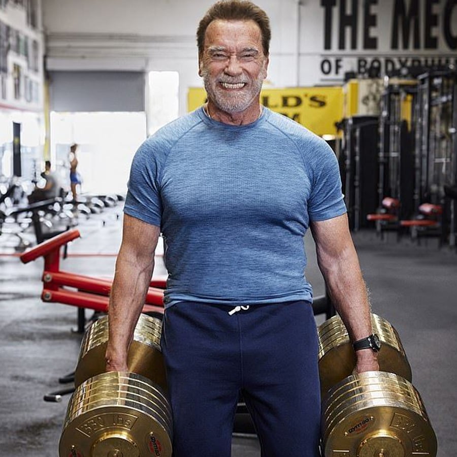 A man in a blue shirt and navy pants stands in a gym, smiling while holding large gold weights in each hand. Gym equipment and wall signs are visible in the background.