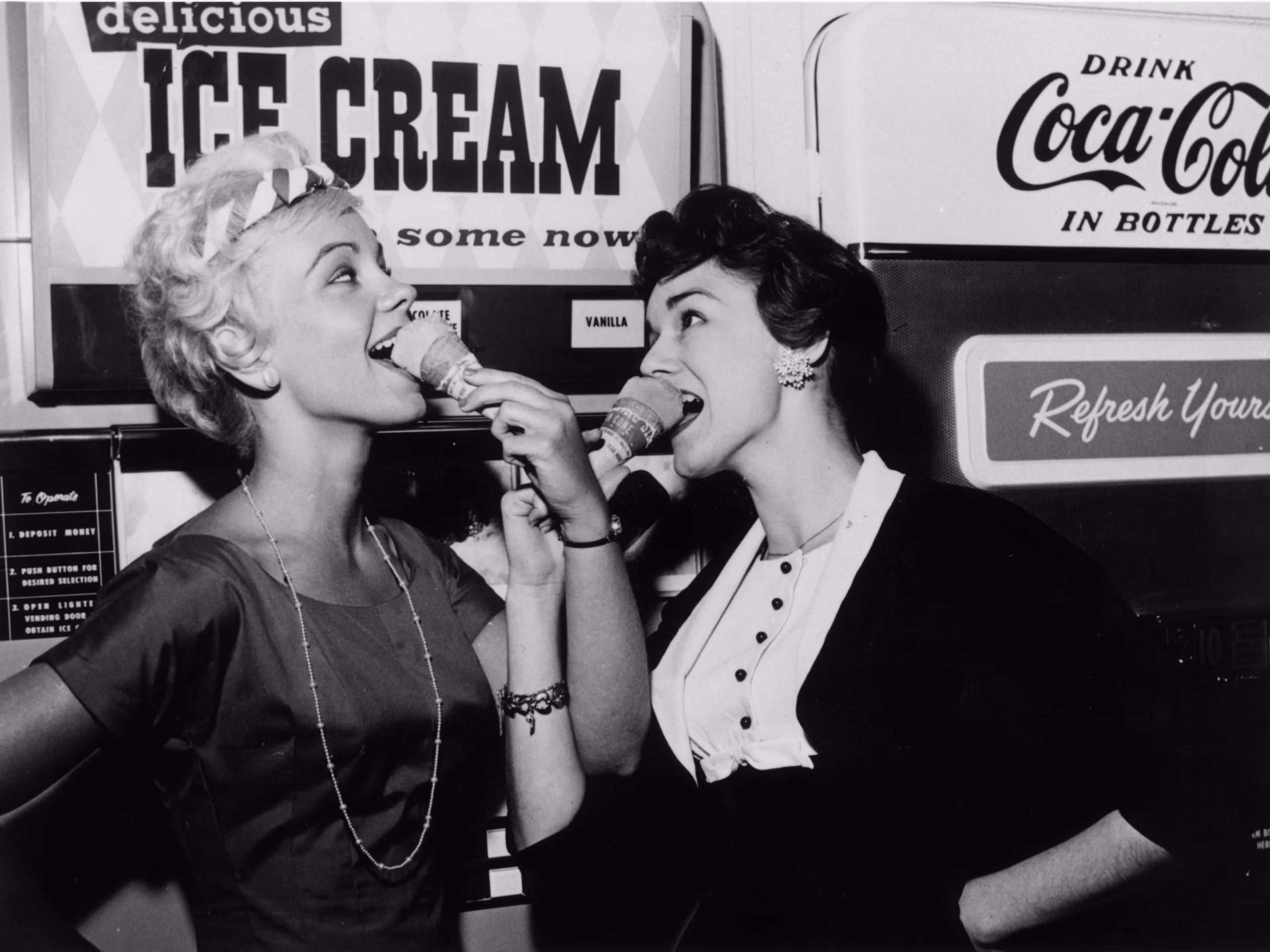 Two women smile and eat ice cream cones in front of vintage signs for ice cream and Coca-Cola, creating a cheerful, retro atmosphere.