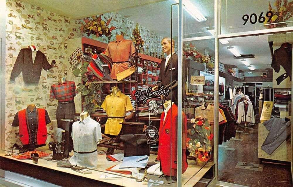 A vintage clothing store display with mannequins dressed in retro shirts, jackets, and ties. Shelves and racks show folded clothes and accessories. The entrance glass door shows the number "9069" above.