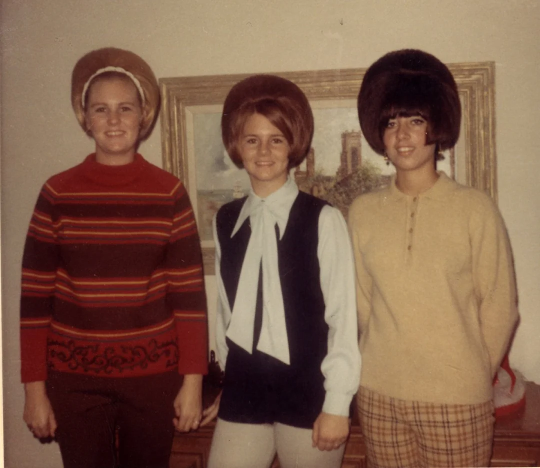 Three women standing indoors in front of a painting, all with voluminous 1960s-style hairdos. They wear vintage clothing: a striped sweater, a tie-neck blouse with vest, and a beige top with plaid pants.