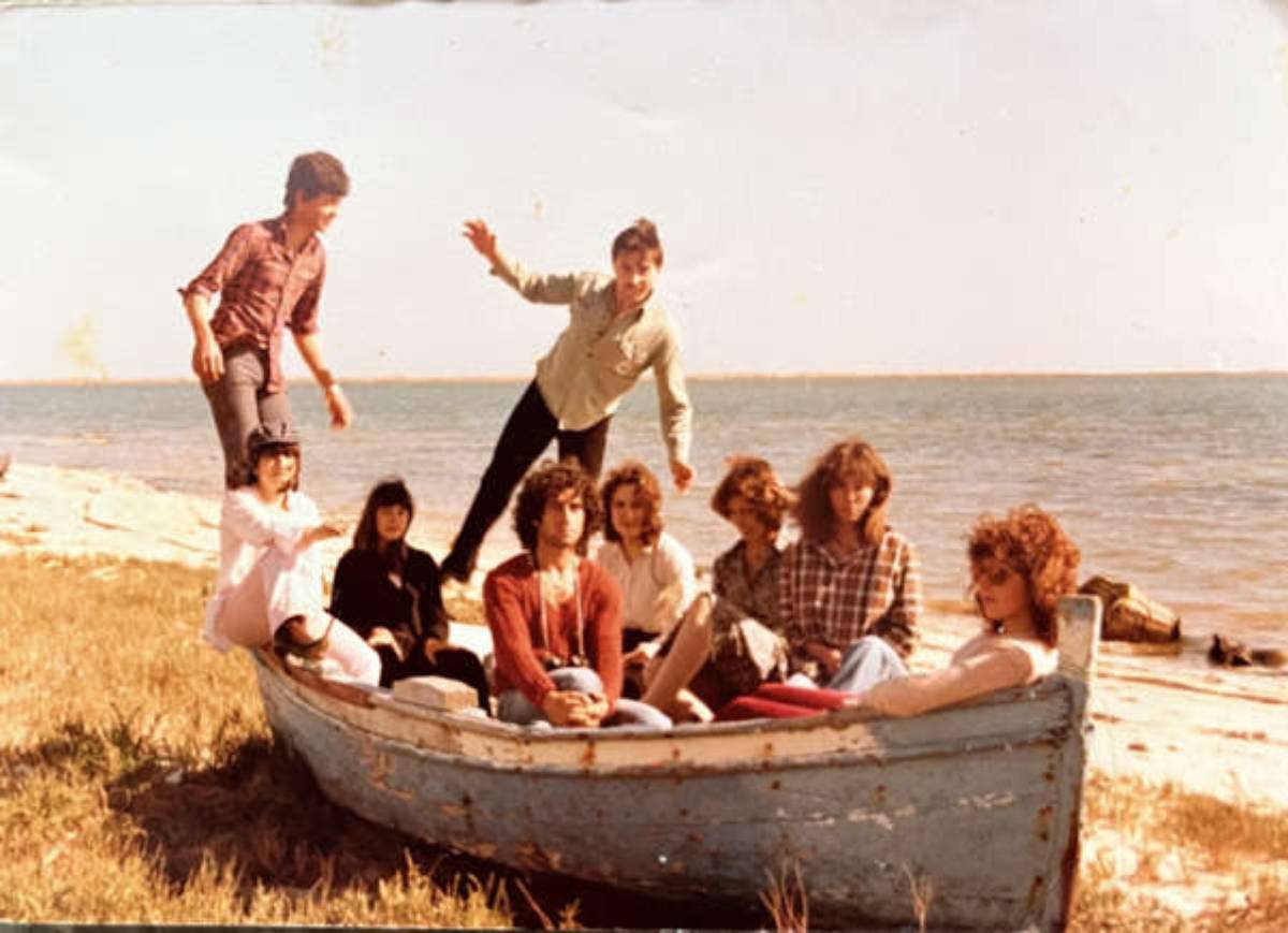 Eight people, some seated inside a weathered rowboat on grass near a shoreline, while others stand or pose playfully behind it. The group appears relaxed, with water and sky in the background.