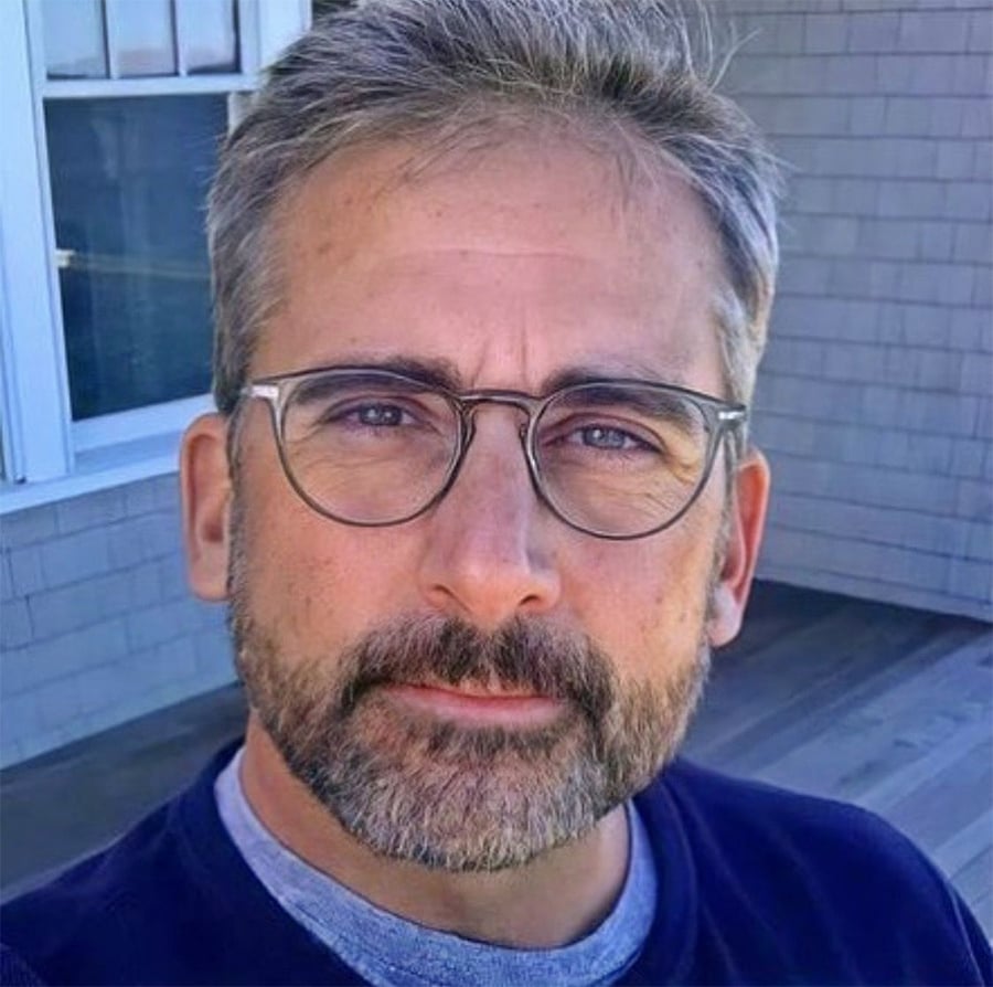 A middle-aged man with gray hair, a beard, and round glasses is standing outside near a window and gray siding, looking directly at the camera with a neutral expression.