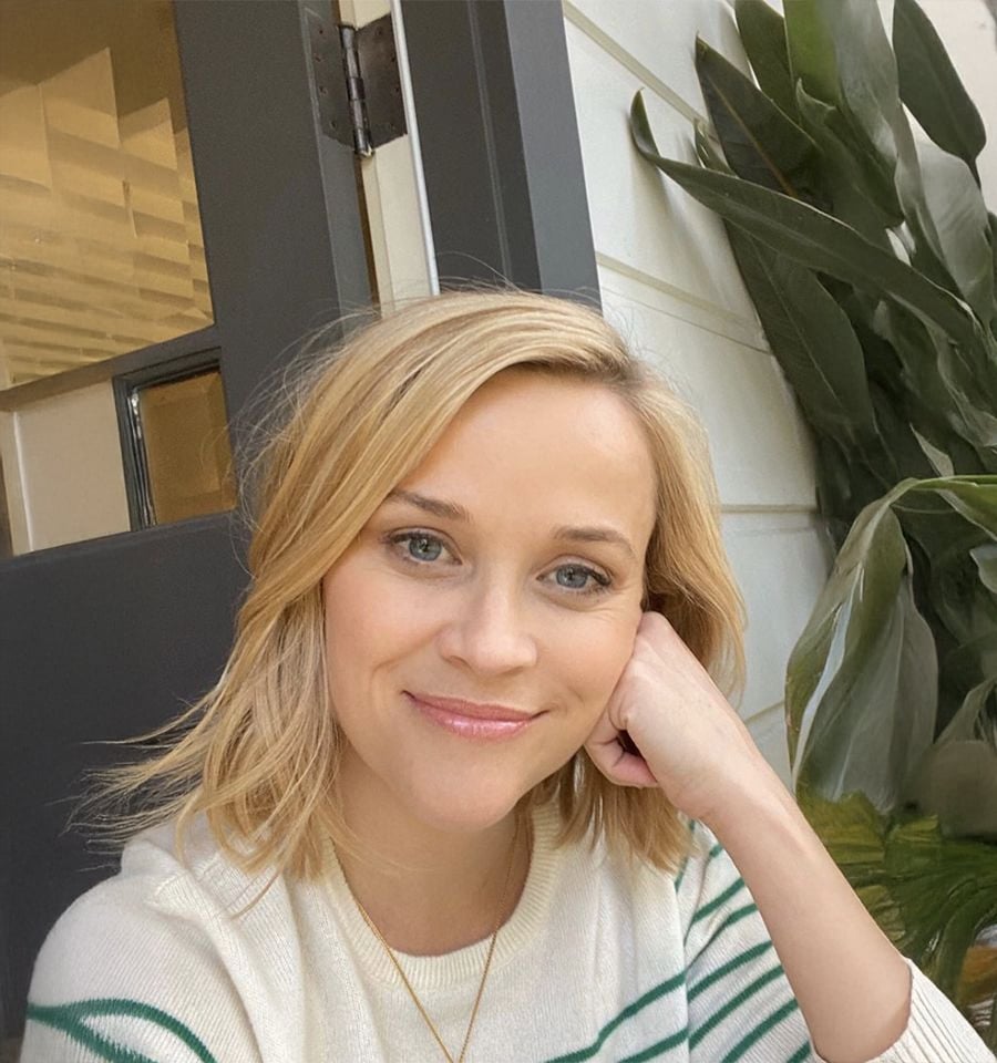 A woman with blonde hair and a light sweater sits indoors, smiling gently, with her head resting on her hand. Green plants and a window are visible in the background.