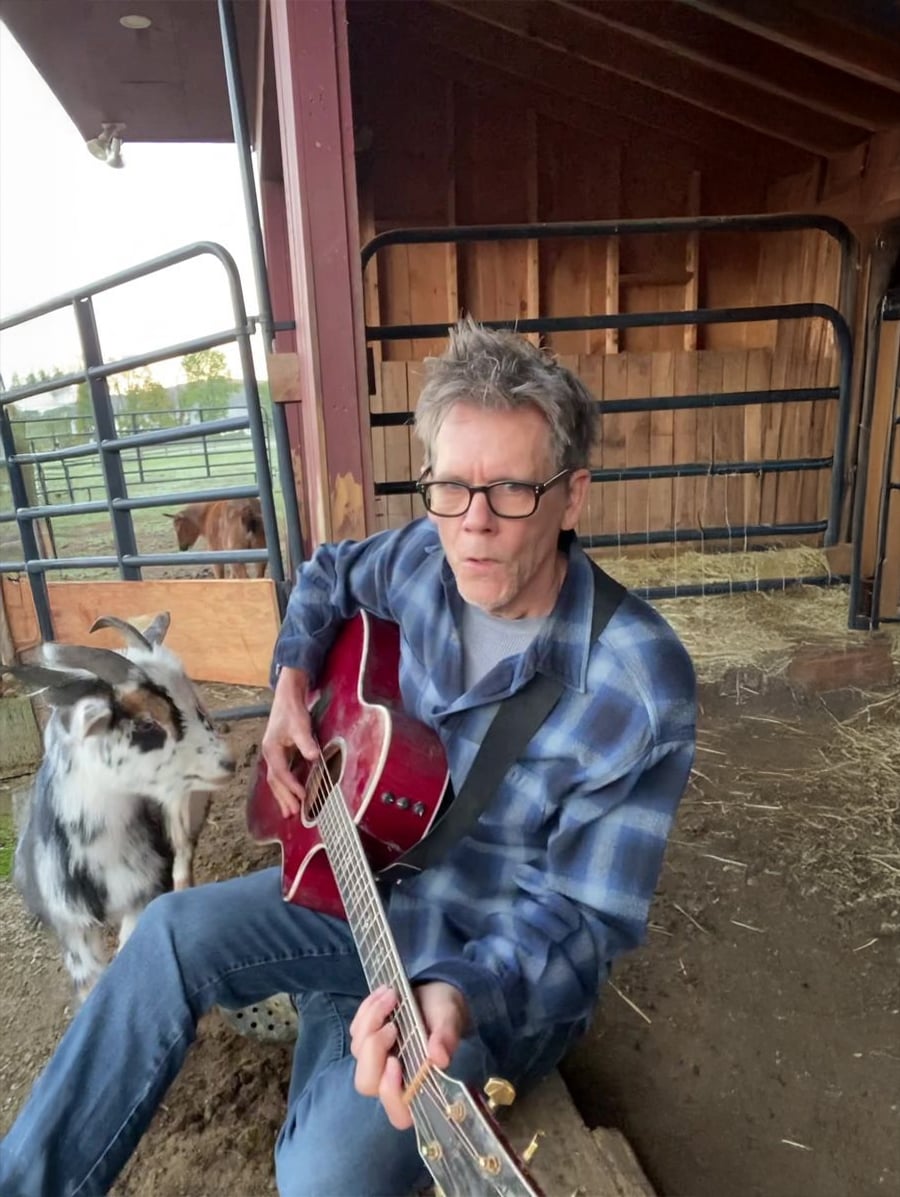 A man wearing glasses and a blue plaid shirt plays a red acoustic guitar while sitting near a goat in a barn with wood walls and outdoor fencing visible in the background.