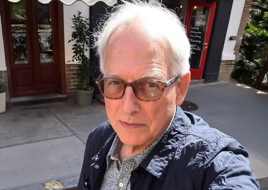 An older man with white hair and glasses takes a selfie outdoors. He is wearing a blue jacket over a patterned shirt, and behind him are building entrances, a red door, and potted plants.