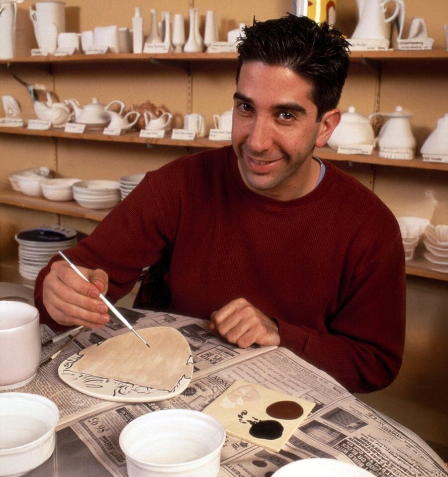 A man with dark hair wearing a red sweater smiles while painting a ceramic plate at a pottery studio, surrounded by shelves filled with various white ceramics and painting supplies.