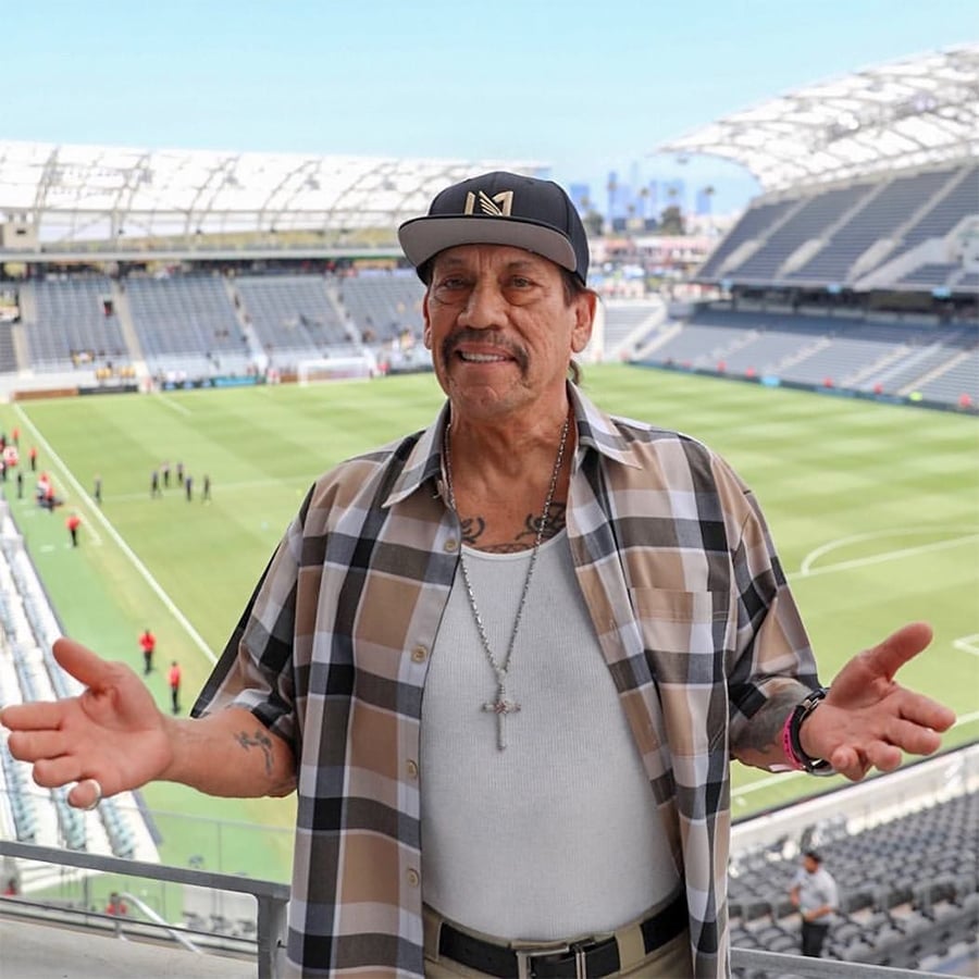 Smiling man in a plaid shirt, tank top, and cap stands in front of a large, empty soccer stadium, arms open and welcoming, with a cross necklace visible.