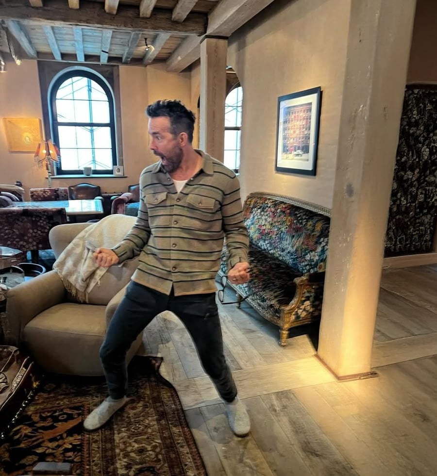 A man in a striped shirt and dark pants stands in a cozy, rustic room with wooden beams, arched windows, patterned furniture, and a lamp. He appears to be excited or dancing, with his fists clenched and mouth open.