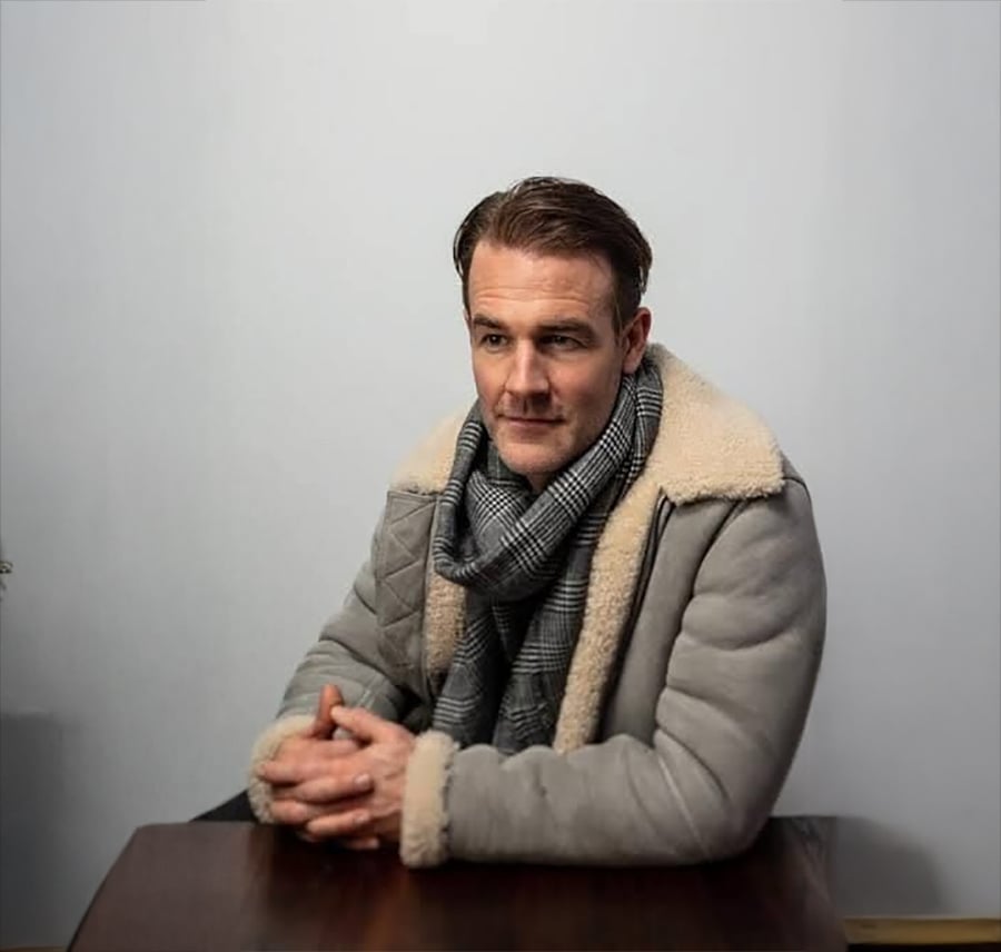 A man sits at a table, wearing a gray shearling coat and a plaid scarf. His hands are clasped, and he looks slightly to the side against a plain, light-colored background.