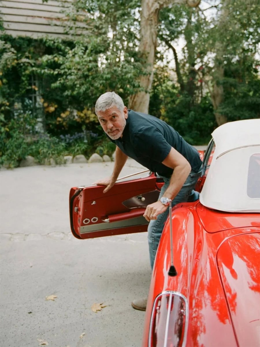A man with gray hair and a beard, wearing a black shirt and jeans, steps out of a classic red convertible car with the door open, in a driveway surrounded by trees and greenery.