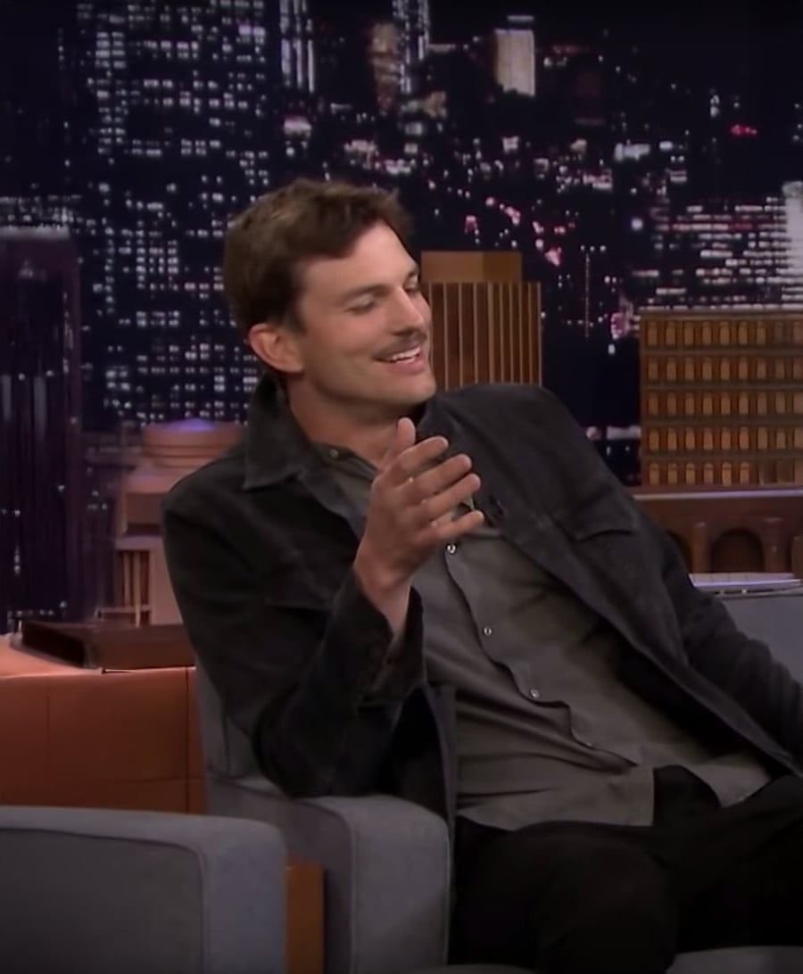 A man with short brown hair, wearing a dark jacket and gray shirt, sits smiling and gesturing with one hand on a talk show set with a cityscape backdrop at night.