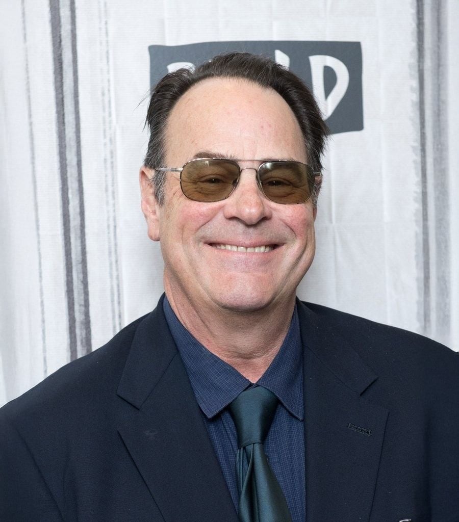 A man wearing sunglasses, a dark suit, blue shirt, and dark tie smiles in front of a light-colored background with partially visible text.