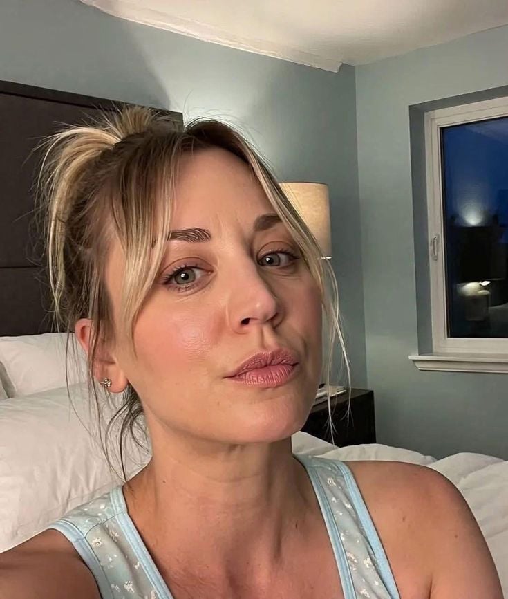 A woman with blonde hair in a messy bun and side-swept bangs poses for a selfie in a softly lit bedroom, wearing a light blue sleeveless top. A bed, lamp, and window are visible in the background.