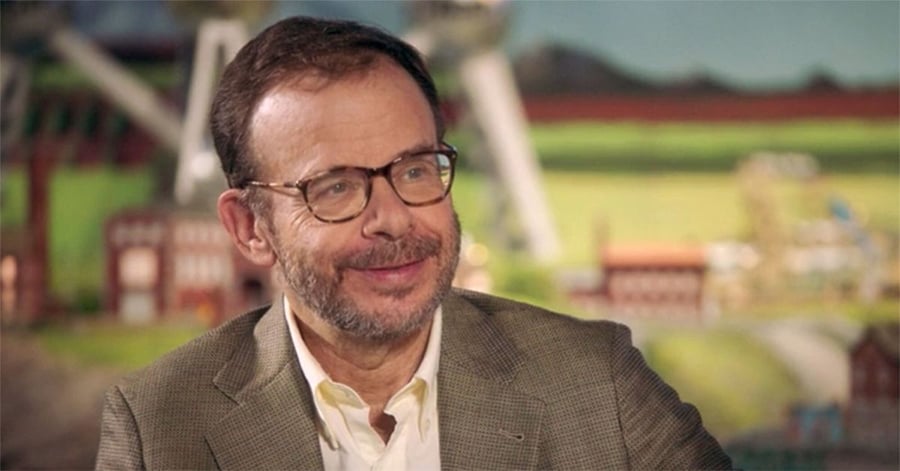 A man in glasses and a brown blazer smiles while sitting indoors. The background is blurred and colorful, possibly depicting a painted or illustrated scene.