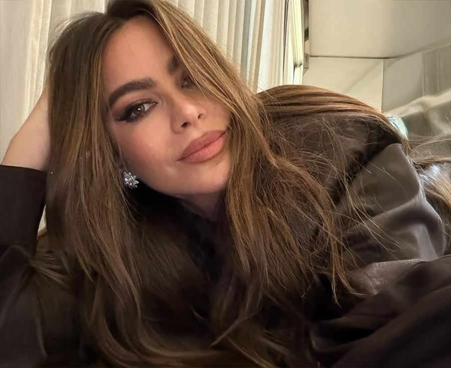 A woman with long brown hair and flawless makeup lies on her side, resting her head on her hand, and looks at the camera. She wears a dark top and large, sparkling earrings. Light curtains are in the background.