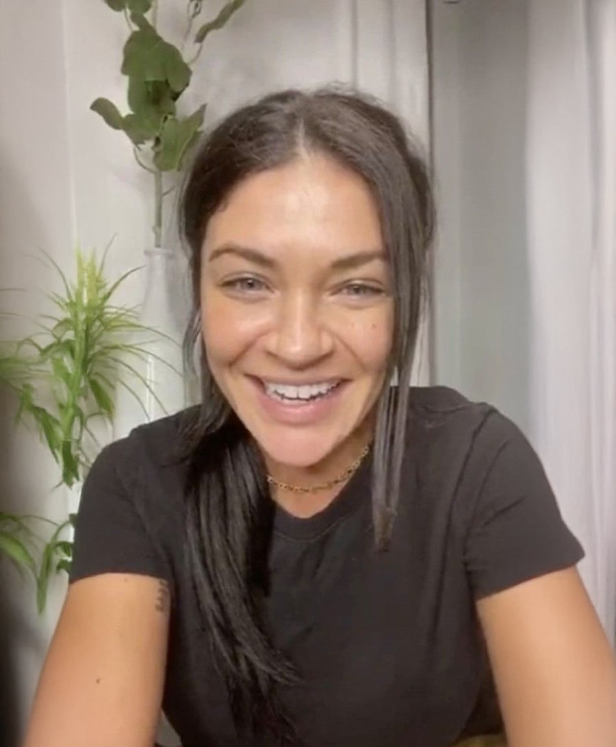 A woman with long dark hair in a ponytail smiles at the camera. She is wearing a black t-shirt and a gold necklace. Green plants are visible in the background.