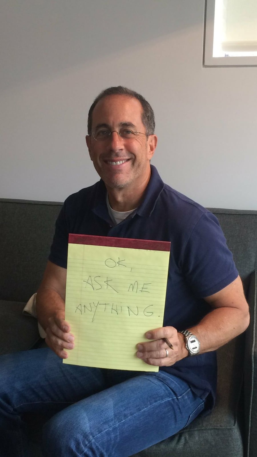 A man sitting on a couch, smiling at the camera, and holding a yellow notepad with the handwritten message "OK ASK ME ANYTHING.