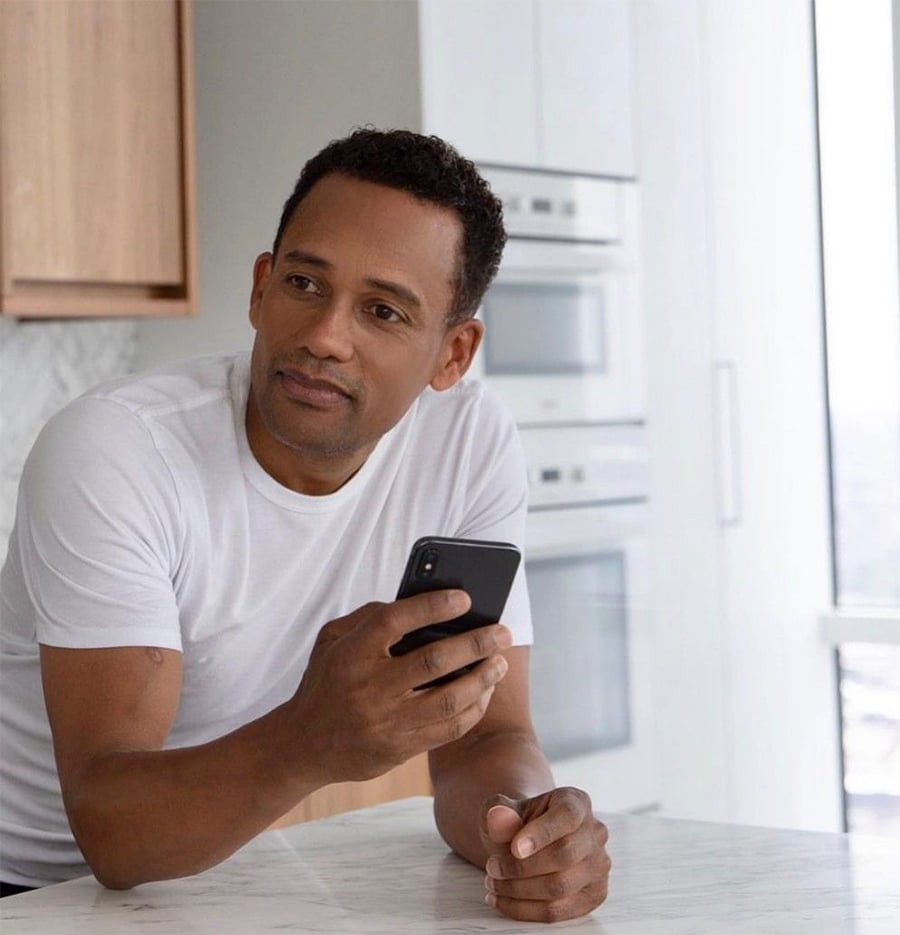 A man in a white t-shirt leans on a kitchen counter, holding a smartphone and looking to the side with a relaxed expression. Modern kitchen appliances are visible in the background.