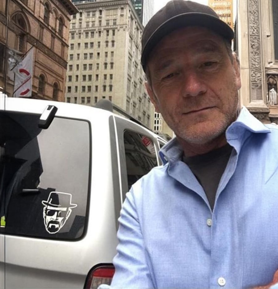 A man in a blue shirt and black cap takes a selfie on a city street next to a white vehicle with a sticker of a bearded, hat-wearing cartoon face on its rear window. Tall buildings are visible in the background.