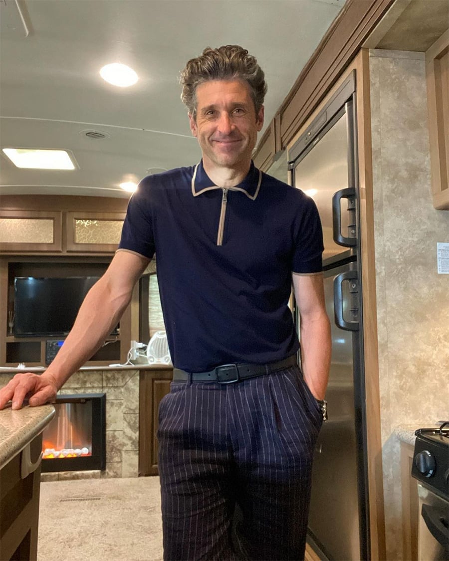 A man with wavy hair, wearing a navy polo shirt and striped pants, stands smiling in a cozy RV kitchen with a fireplace, TV, and wooden cabinets in the background.