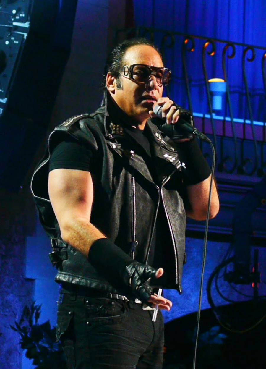 A man wearing sunglasses, fingerless gloves, and a black leather vest holds a microphone and performs on stage, with a blue-lit background and a balcony railing visible behind him.