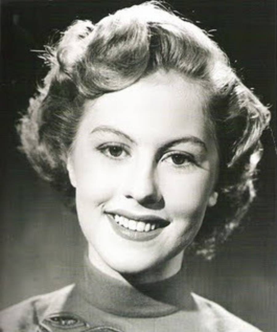 Black and white portrait of a smiling young woman with short, wavy hair, wearing a high-collared top. The background is plain and softly lit.
