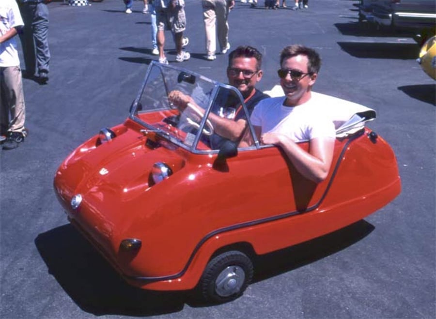 Two smiling men are seated in a very small, red, vintage three-wheeled car at an outdoor event. Other people and vehicles are visible in the background on a sunny day.