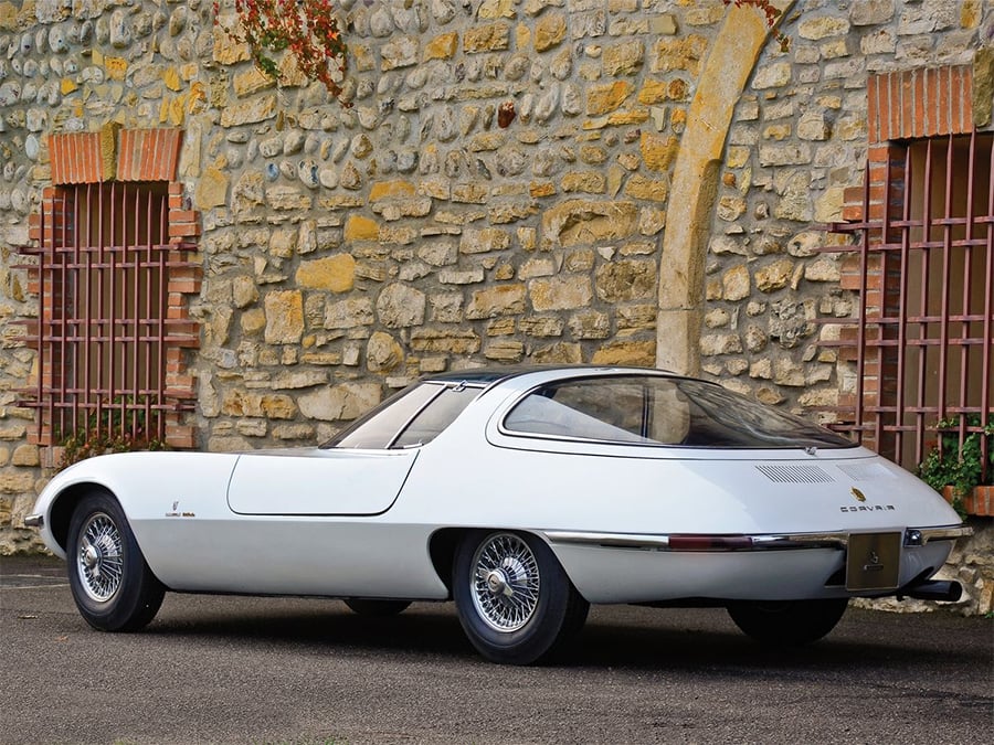 A vintage white sports car with a sleek, futuristic design is parked in front of a rustic stone wall with barred windows and climbing plants.