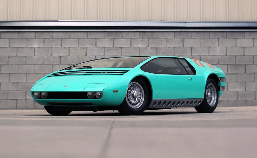 A turquoise vintage sports car with a low, wide body, large wheels, and black stripe details, parked on pavement in front of a concrete block wall and tan building.