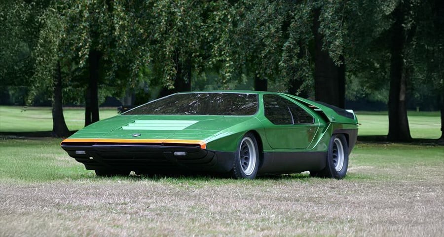 A green, futuristic sports car with a wedge-shaped design is parked on grass, surrounded by trees in the background.