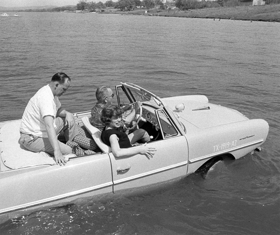Three people ride in a white amphibious car driving through water near a shore, with the driver smiling and one passenger sitting on the back.