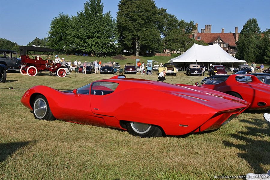 A sleek, bright red concept car with smooth curves is displayed on a grassy field with other classic cars, people, a large tent, and trees in the background.