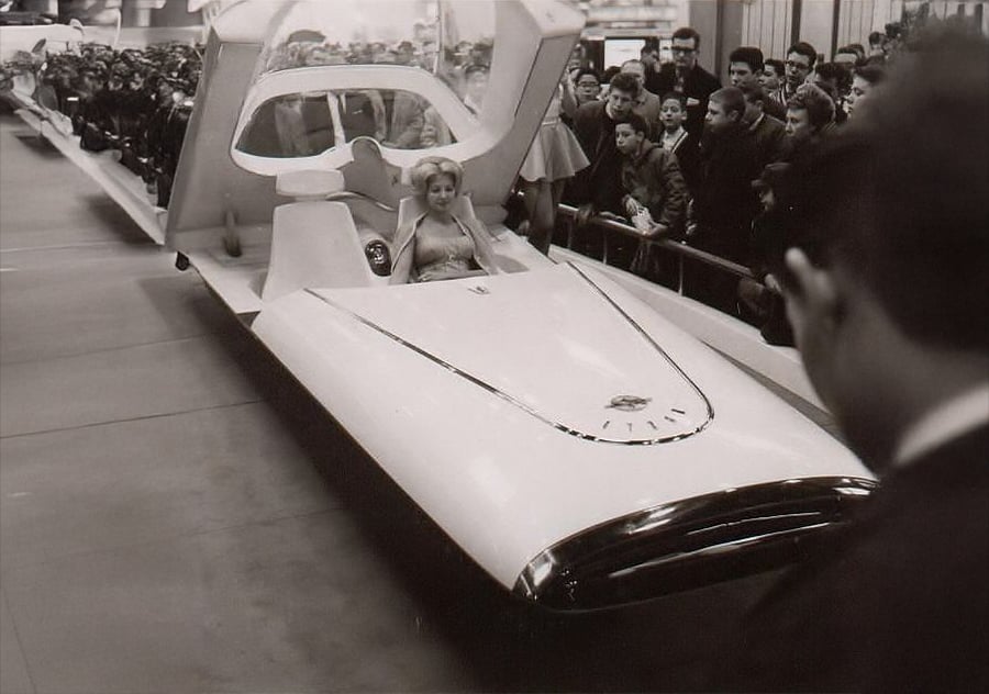 A futuristic car with an open canopy is displayed at an exhibition. A woman sits inside, while a crowd of onlookers surrounds the vehicle, watching with interest. The photo is black and white.