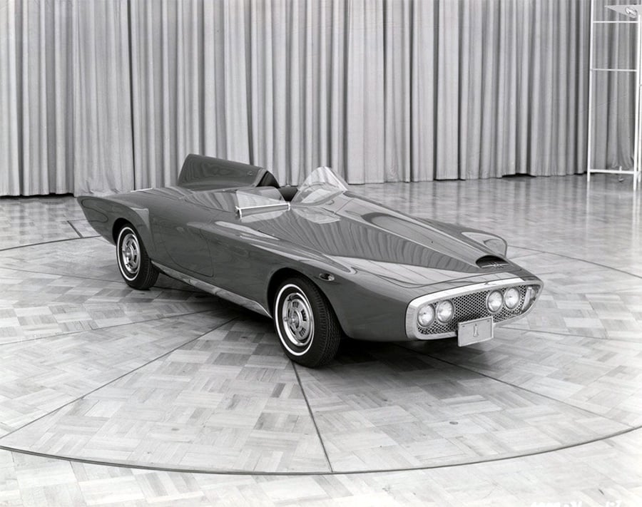 A sleek, vintage concept car with a low profile, dual windshields, and prominent front grille is displayed indoors on a circular wood floor with curtains in the background.