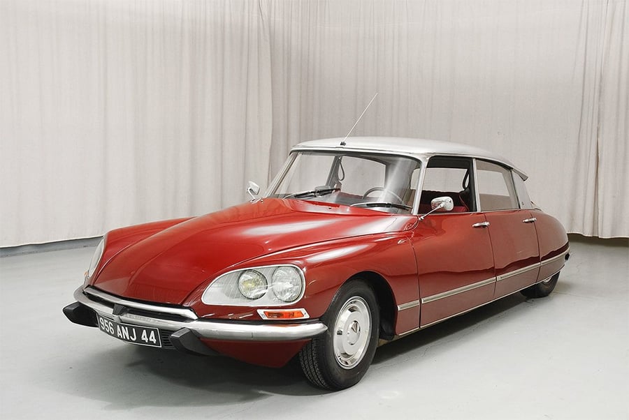 A classic red Citroën DS car with a silver roof is parked indoors on a light-colored floor, in front of a plain white curtain backdrop.
