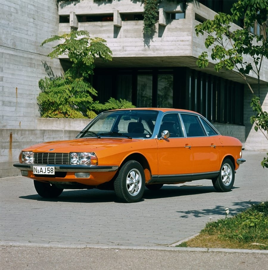 An orange classic sedan car is parked on a stone driveway in front of a modern concrete building, with green plants and a small tree nearby.