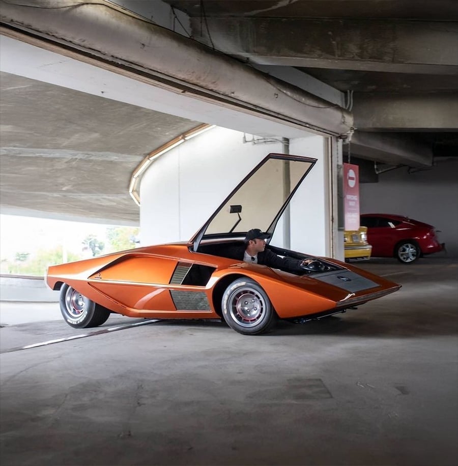 An orange wedge-shaped sports car with a gullwing door open is parked in a concrete parking garage. A person is seated inside the car.