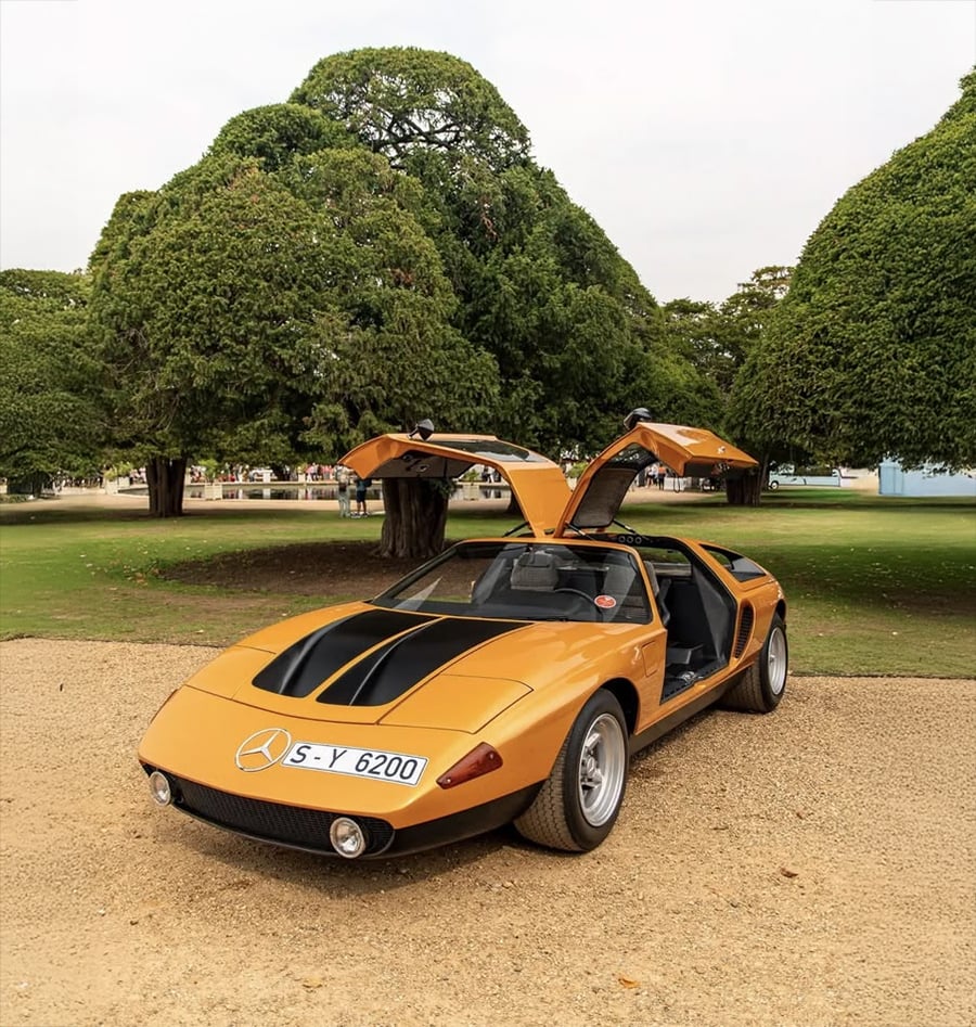 A vintage orange sports car with gullwing doors open is parked on a gravel path, with large green trees in the background.