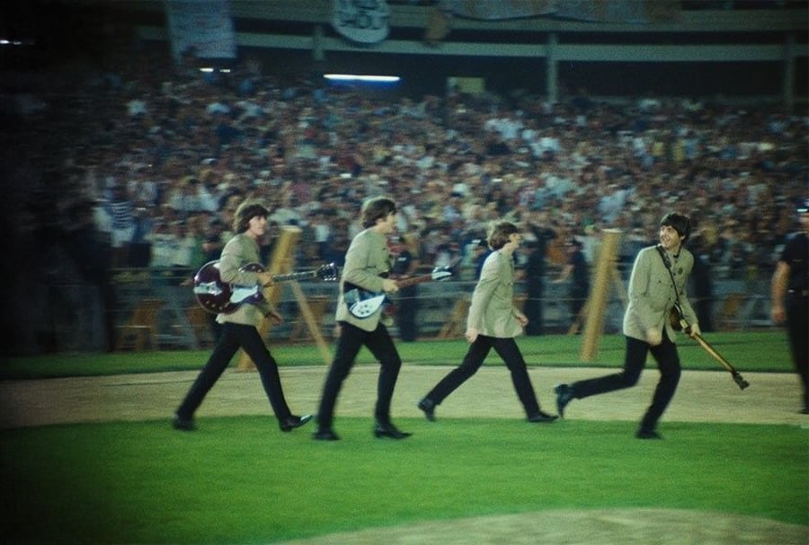 Four musicians in matching jackets walk across a field carrying guitars, with a large crowd of cheering fans in a stadium behind them. The scene appears lively and energetic.