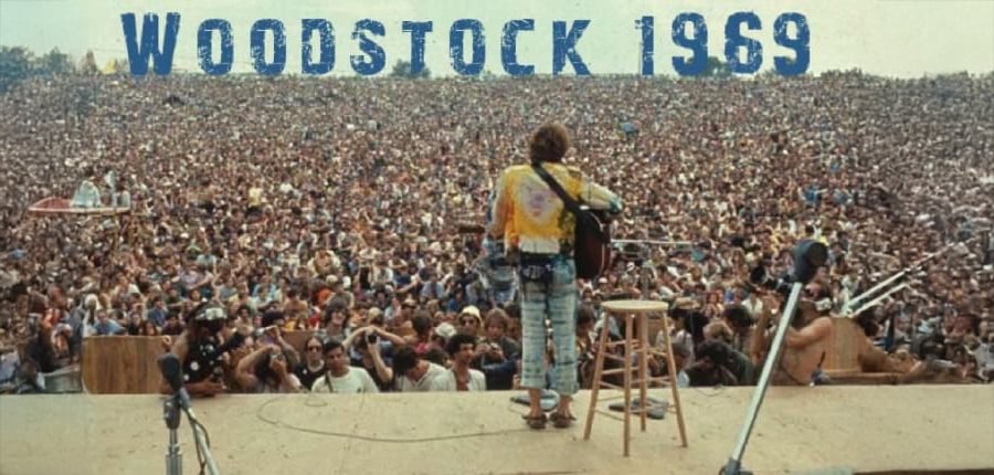A musician stands on stage with a guitar, facing a massive crowd at Woodstock 1969. The audience stretches into the distance, and "WOODSTOCK 1969" is written in large blue letters at the top of the image.