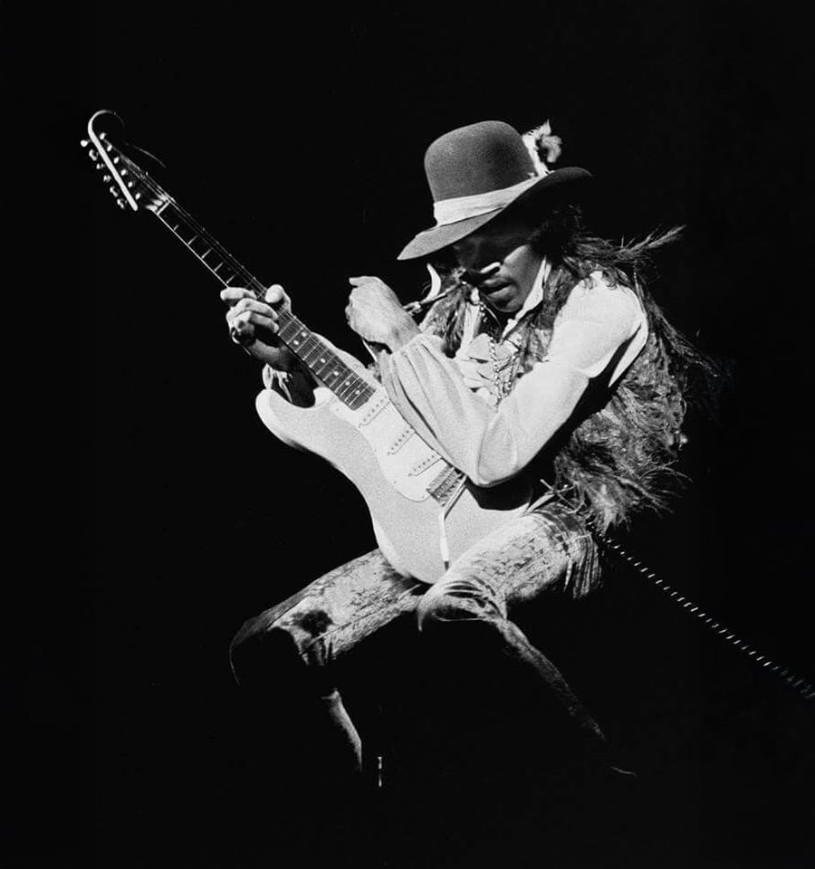 A musician wearing a wide-brimmed hat and feathered vest plays an electric guitar energetically on stage against a dark background.