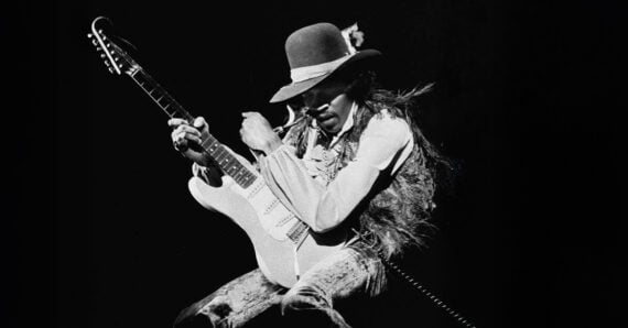 A musician wearing a hat and feathered vest plays an electric guitar energetically on stage against a dark background.
