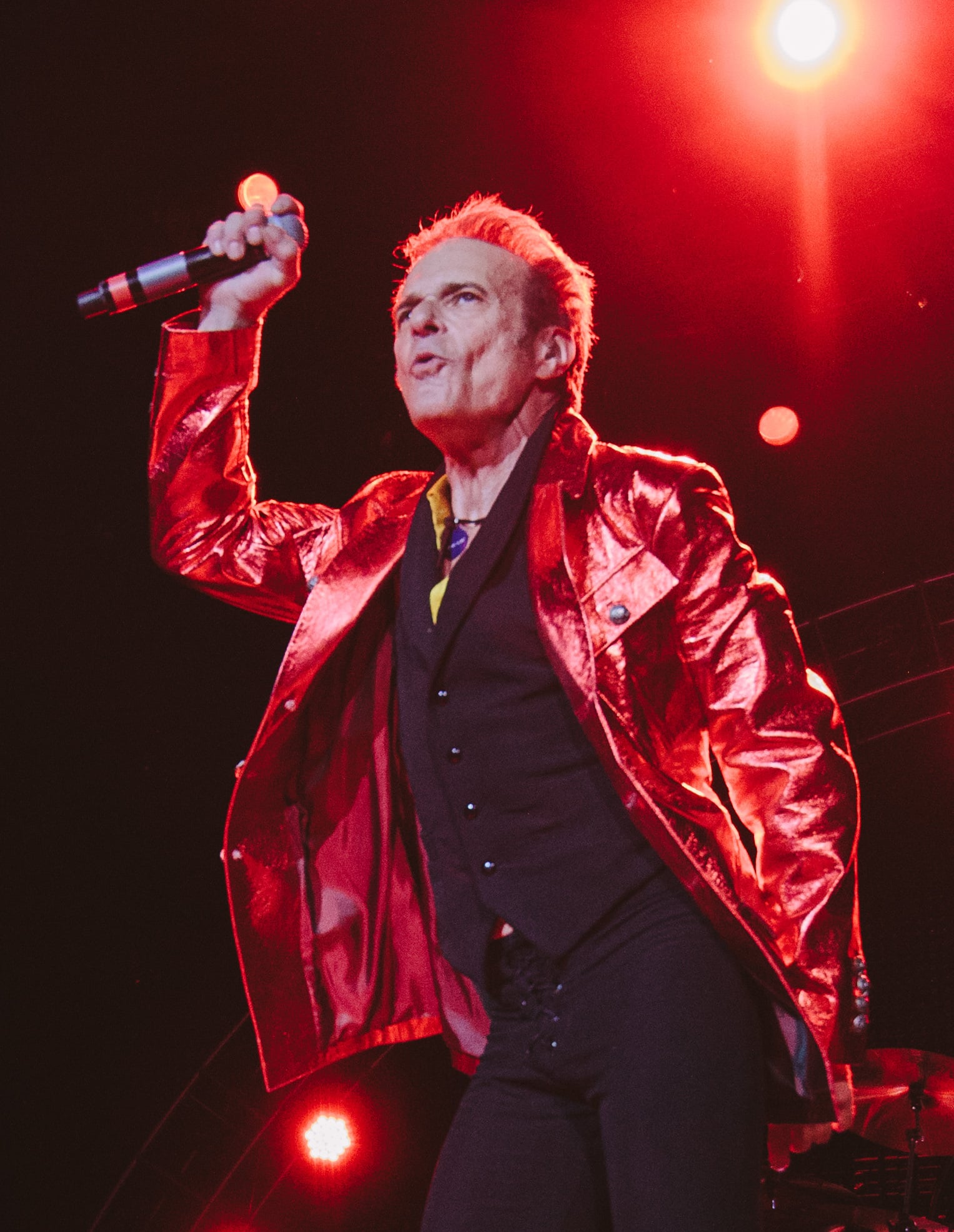 A singer in a shiny red jacket holds a microphone and performs energetically on stage under bright red lights.